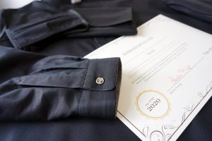 Detailaufnahme einer Anfertigung, explizit: ein Herrenhemd in schwarz; außerdem ist ein Ausschnitt eines Anfertigungszertifikats zu sehen.